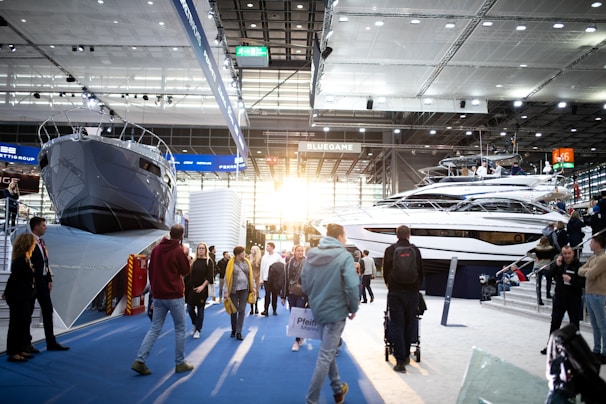 A boat exhibition featuring large, luxury yachts indoors, with several people walking around and observing. The setting includes a modern, well-lit space with high ceilings and large windows letting in natural light.