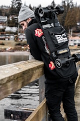 A person in a gray beanie and black clothing is leaning against a wooden railing, overlooking a water body. They have an electric skateboard strapped to their back with the brand 'Basecamp' visible. In the background, there are trees and houses on a hillside, giving the setting an outdoor, serene vibe.