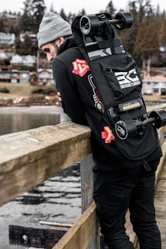 A person in a gray beanie and black clothing is leaning against a wooden railing, overlooking a water body. They have an electric skateboard strapped to their back with the brand 'Basecamp' visible. In the background, there are trees and houses on a hillside, giving the setting an outdoor, serene vibe.