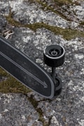A skateboard with black wheels and a dark deck, featuring a white arrow design, is placed on a rough, textured surface covered with patches of green moss and gray stones.