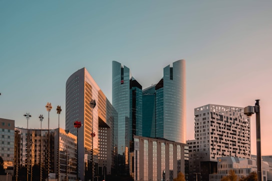 A modern cityscape featuring several tall, uniquely designed skyscrapers with glass facades under a clear sky. The buildings capture the warm glow of the setting sun, adding a gleam to their surfaces. Some architectural elements stand out with distinct patterns and shapes. Street lamps and a few trees are also visible in the foreground.