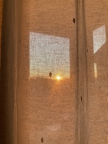 A textured linen curtain partially drawn over a window with warm sunset light spilling through, highlighting a plush cushion on the sill.