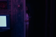 A cinematic shot of a dark room illuminated only by the glow of a TV screen showing an intense drama.
