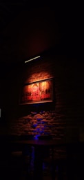 A dimly lit interior with a brick wall forms the backdrop. Centered is a framed picture or poster illuminated by red and orange lighting, casting a warm glow. Below the frame, a dark wooden table and chair are visible in the shadows. Blue lighting adds a subtle accent from beneath.