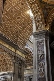 In the interior of an ornate cathedral, stunning architecture is highlighted with intricate patterns on the arched ceiling. Gold and marble elements are predominant, embodying Renaissance artistry. Latin inscriptions and decorative sculptures of cherubs and figures are prominently displayed on the walls and columns.