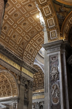 In the interior of an ornate cathedral, stunning architecture is highlighted with intricate patterns on the arched ceiling. Gold and marble elements are predominant, embodying Renaissance artistry. Latin inscriptions and decorative sculptures of cherubs and figures are prominently displayed on the walls and columns.