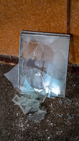 A broken mirror with fragments scattered on the floor, set against a textured brown wall. The glass shards reflect light, creating a subtle contrast with the dark carpet.