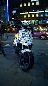 A scooter parked on a city street is covered with numerous colorful stickers. It is situated on a paved sidewalk, and there is another bicycle partially visible next to it. The backdrop features a building with illuminated windows and neon signs, characteristic of an urban night setting.