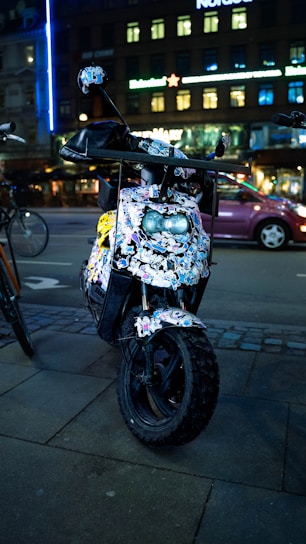 A scooter parked on a city street is covered with numerous colorful stickers. It is situated on a paved sidewalk, and there is another bicycle partially visible next to it. The backdrop features a building with illuminated windows and neon signs, characteristic of an urban night setting.