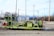 A heavy rail maintenance vehicle stands on the tracks, painted in green with various mechanical components visible. The background includes industrial buildings, power lines, and poles, with a mostly cloudy sky overhead.