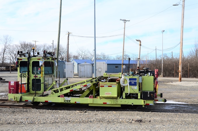 A heavy rail maintenance vehicle stands on the tracks, painted in green with various mechanical components visible. The background includes industrial buildings, power lines, and poles, with a mostly cloudy sky overhead.
