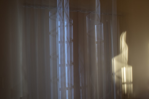 Soft blurred vertical lines and abstract shapes create a gentle, dreamy atmosphere. Light blue and beige tones prevail, with subtle hints of shadow and light interplay.
