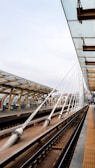 A modern train station platform with an overhead glass canopy supported by wooden beams and white steel cables. The platform features clean lines and a geometric design. Multiple tracks are visible, showing a well-maintained infrastructure. City buildings are seen in the background, and the sky is cloudy.