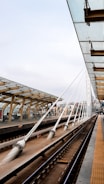 A modern train station platform with an overhead glass canopy supported by wooden beams and white steel cables. The platform features clean lines and a geometric design. Multiple tracks are visible, showing a well-maintained infrastructure. City buildings are seen in the background, and the sky is cloudy.