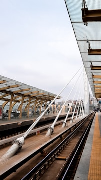 A modern train station platform with an overhead glass canopy supported by wooden beams and white steel cables. The platform features clean lines and a geometric design. Multiple tracks are visible, showing a well-maintained infrastructure. City buildings are seen in the background, and the sky is cloudy.