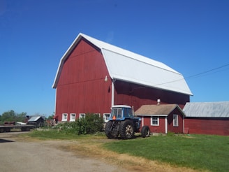 A large red barn with a white roof stands prominently against a clear blue sky. In front of the barn, a blue tractor is parked on a patch of grass. Surrounding the barn are smaller red outbuildings and some greenery, indicating a rural farm setting.