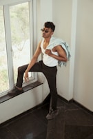 A person wearing sunglasses and a white tank top is standing near a large window. They are holding a light denim jacket over one shoulder while resting one foot on the window ledge. The person looks relaxed and confident, with a modern and casual style.