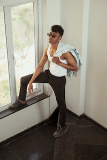 A person wearing sunglasses and a white tank top is standing near a large window. They are holding a light denim jacket over one shoulder while resting one foot on the window ledge. The person looks relaxed and confident, with a modern and casual style.