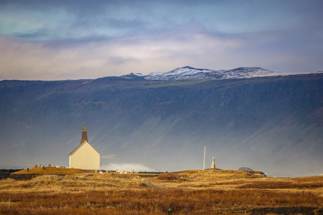 An isolated white church with a brown roof is situated in a vast open field of tall, dry grass. In the background, a large mountain range with patches of snow on its peaks can be seen under a cloudy sky. The field surrounding the church appears peaceful and expansive, with minimal human activity visible.