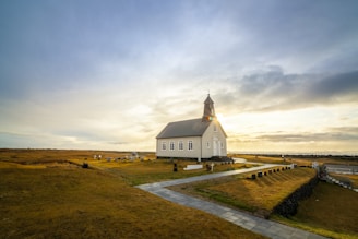 A small, charming church with a steeple is situated in a vast open field. The building is painted white and contrasts with the golden hues of the surrounding grassland. The sky is partly cloudy with sunlight breaking through, casting a warm glow over the scene. In the foreground, a walkway leads up to the church, and a cemetery with several headstones is visible.