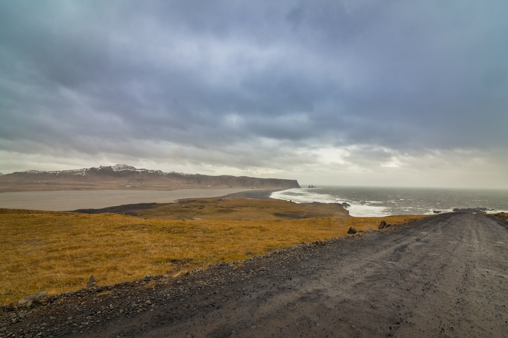 a dirt road near a body of water under a cloudy sky