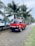 A red mini truck is parked on a road lined with palm trees. The vehicle has Japanese characters on it and a license plate reading JBY 4019. The background shows a cloudy sky and more parked vehicles along the road.