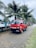 A red mini truck is parked on a road lined with palm trees. The vehicle has Japanese characters on it and a license plate reading JBY 4019. The background shows a cloudy sky and more parked vehicles along the road.