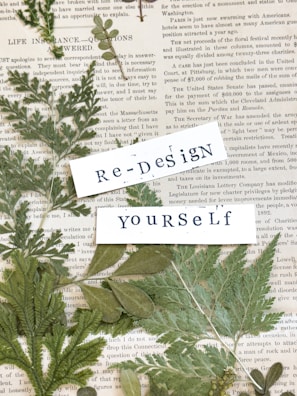 An arrangement of dried green leaves placed on top of vintage newspaper pages. Overlaying the leaves are two pieces of white paper with the words 'Re-Design' and 'Yourself' printed in a typewriter font.
