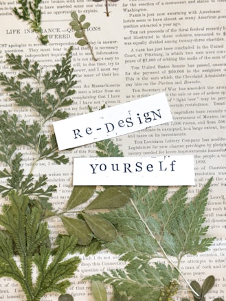 An arrangement of dried green leaves placed on top of vintage newspaper pages. Overlaying the leaves are two pieces of white paper with the words 'Re-Design' and 'Yourself' printed in a typewriter font.