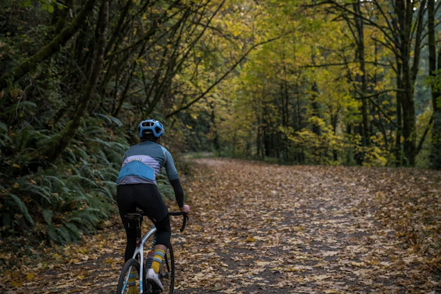 A cyclist riding a gravel bike on a sunlit forest trail with autumn leaves scattered around.