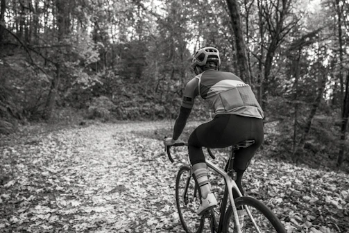 A cyclist riding a gravel bike on a sunlit forest trail with autumn leaves.