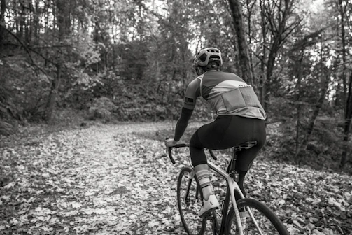 A cyclist riding a gravel bike on a sunlit forest trail scattered with autumn leaves.