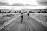 Two cyclists ride along a gravel road that stretches into the distance, flanked by open fields and farmhouses. The sky is cloudy, adding a dramatic backdrop to the rural landscape.