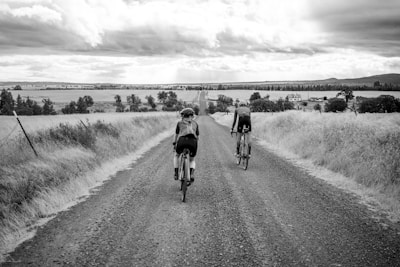 Two cyclists ride along a gravel road that stretches into the distance, flanked by open fields and farmhouses. The sky is cloudy, adding a dramatic backdrop to the rural landscape.