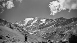 A lone hiker with a backpack and trekking poles walks across a rocky and rugged terrain towards a majestic snow-capped mountain range. The landscape is vast and awe-inspiring, with dramatic cloud formations in the sky.