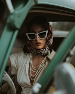 A woman sits in an old-fashioned car, exuding a sense of vintage glamour. She wears large, white cat-eye sunglasses and a colorful scarf tied around her head. Her outfit includes a white blouse and layered pearl necklaces. The mood is stylish and reminiscent of classic Hollywood.