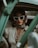 A woman sits in an old-fashioned car, exuding a sense of vintage glamour. She wears large, white cat-eye sunglasses and a colorful scarf tied around her head. Her outfit includes a white blouse and layered pearl necklaces. The mood is stylish and reminiscent of classic Hollywood.