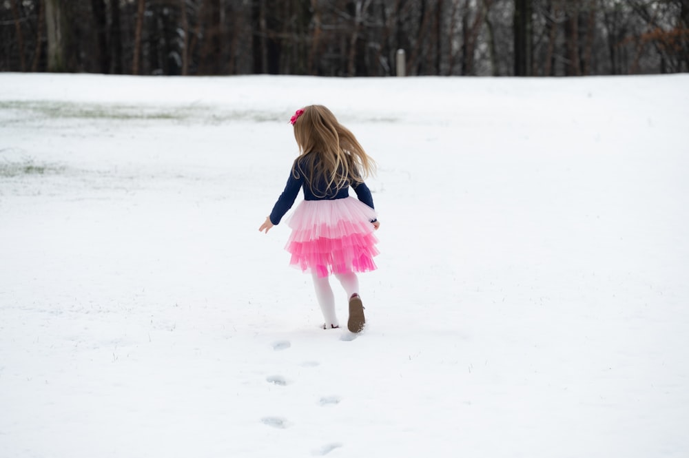 a little girl in a pink and blue dress walking in the snow
