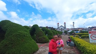 A man holding a child in a park-like setting with large, sculpted bushes and a historic church with twin towers in the background. There is a sign in Spanish indicating smoking is prohibited and several people are present in the area.