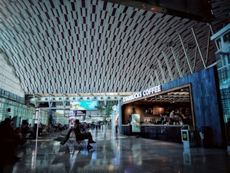A spacious airport terminal with a high, intricately designed ceiling and large windows allowing natural light. Passengers sit in chairs waiting, while a Starbucks Coffee shop is visible on the right side.