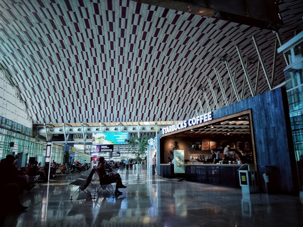 A spacious airport terminal with a high, intricately designed ceiling and large windows allowing natural light. Passengers sit in chairs waiting, while a Starbucks Coffee shop is visible on the right side.