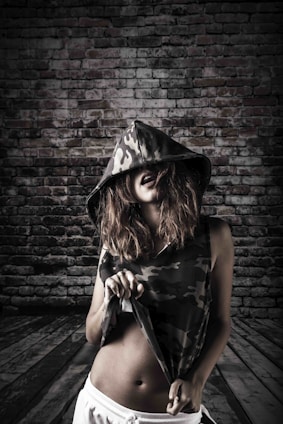 A person with long hair wears a camo-patterned sleeveless hoodie, partially concealing their face with the hood. They hold up the bottom of their top with one hand, revealing their midriff. The background is a rustic brick wall and wooden floor, creating an urban and edgy setting.