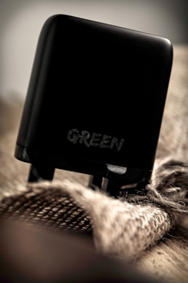 A black charging adapter with the word 'GREEN' written on it is placed on a textured, burlap-like fabric. The lighting is soft, creating a moody and subdued ambiance.