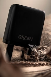 A close-up view of a black electrical plug with the word 'GREEN' engraved on it. The plug is resting on a textured, burlap-like surface, with frayed rope visible nearby. The lighting is soft, highlighting the smooth texture of the plug.