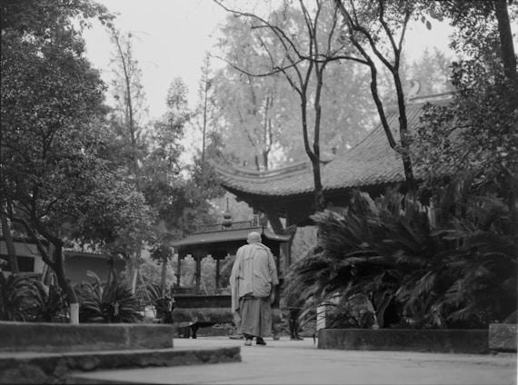A serene outdoor scene featuring a person in traditional attire standing in a garden with lush greenery and an ornate pagoda-style structure in the background. Tall trees surround the courtyard, adding to the tranquil atmosphere.