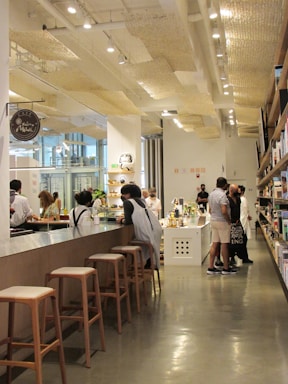 A modern cafe environment with people interacting at a long countertop. There are wooden stools along the counter, and shelves filled with books or items on the right. The cafe has a high ceiling with stylish lighting fixtures and a concrete floor, offering a contemporary and cozy atmosphere.