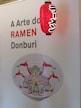 Poster featuring the text 'A Arte do RAMEN Donburi' with a red lantern decorated in Japanese style hanging next to it. Below the text, there is a bowl with a colorful design of a web pattern and intricate face art.