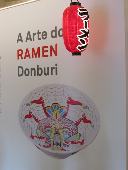Poster featuring the text 'A Arte do RAMEN Donburi' with a red lantern decorated in Japanese style hanging next to it. Below the text, there is a bowl with a colorful design of a web pattern and intricate face art.