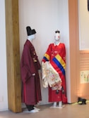 Two mannequins are dressed in traditional Korean attire. The mannequin on the left wears a dark red hanbok with a black headpiece. The mannequin on the right is adorned in a vibrant red garment with a multicolored sash and detailed floral embroidery. Both are positioned against a white wall with a wooden beam and an informational display in the background.