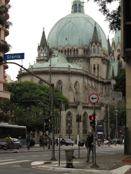 A large, historical cathedral with a green dome is seen from a street intersection. The cathedral features Gothic-style architecture with intricate details. There are street signs, traffic lights, a bus, a cyclist, a pedestrian, and trees in the foreground. The prominent blue street sign reads 'Glória,' and there are buildings in the background.